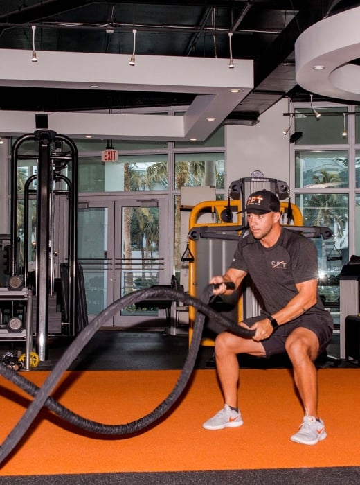 A man in a "GET CPT" hat and "CPT" shirt squats, performing battle rope exercises on orange and black gym flooring. Text visible: "BOXING", "EXIT", "Life Fitness", "NordicTrack", "CLARITY FITNESS COACHING TRAINING", "LYNX".