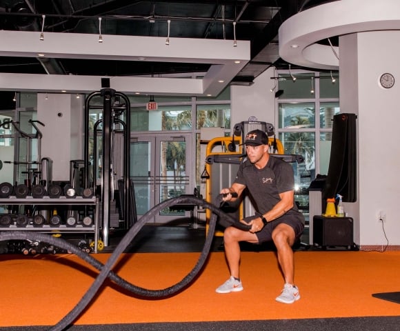 A man in a "GET CPT" hat and "CPT" shirt squats, performing battle rope exercises on orange and black gym flooring. Text visible: "BOXING", "EXIT", "Life Fitness", "NordicTrack", "CLARITY FITNESS COACHING TRAINING", "LYNX".