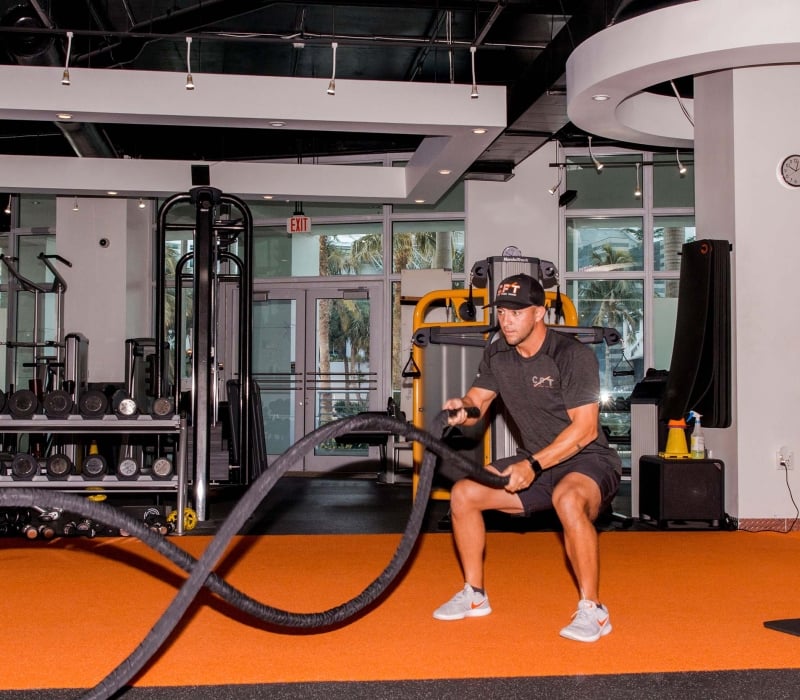 A man in a "GET CPT" hat and "CPT" shirt squats, performing battle rope exercises on orange and black gym flooring. Text visible: "BOXING", "EXIT", "Life Fitness", "NordicTrack", "CLARITY FITNESS COACHING TRAINING", "LYNX".