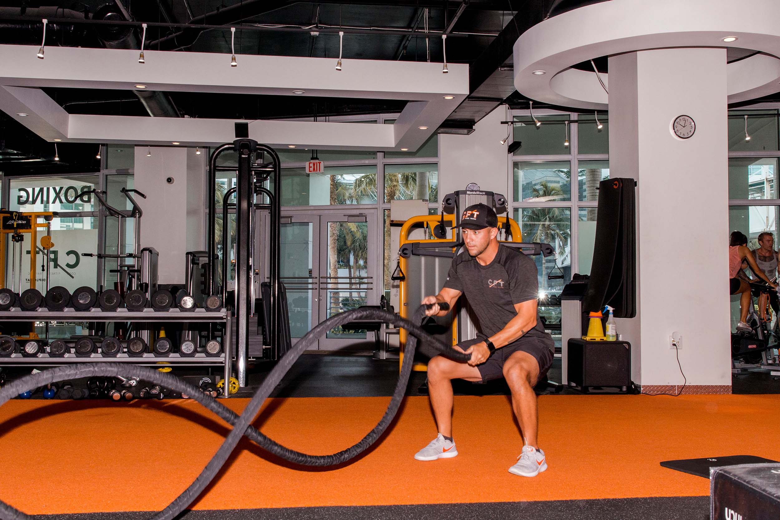 A man in a "GET CPT" hat and "CPT" shirt squats, performing battle rope exercises on orange and black gym flooring. Text visible: "BOXING", "EXIT", "Life Fitness", "NordicTrack", "CLARITY FITNESS COACHING TRAINING", "LYNX".