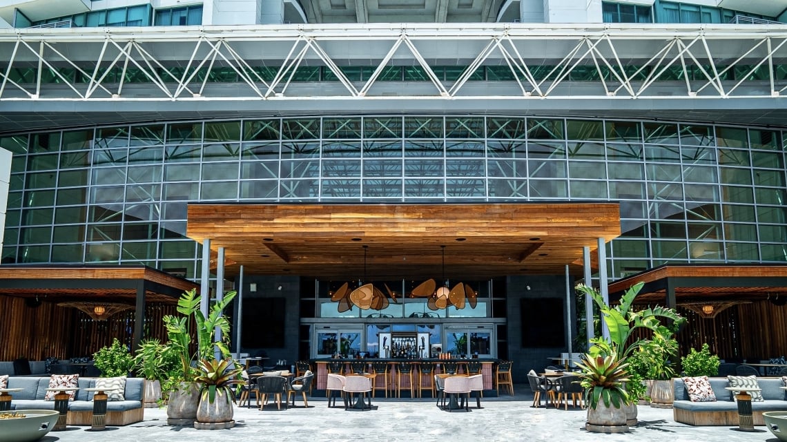 A modern outdoor bar and lounge area is situated beneath a multi-story glass facade, with a high-rise building visible above, connected by a white truss bridge.