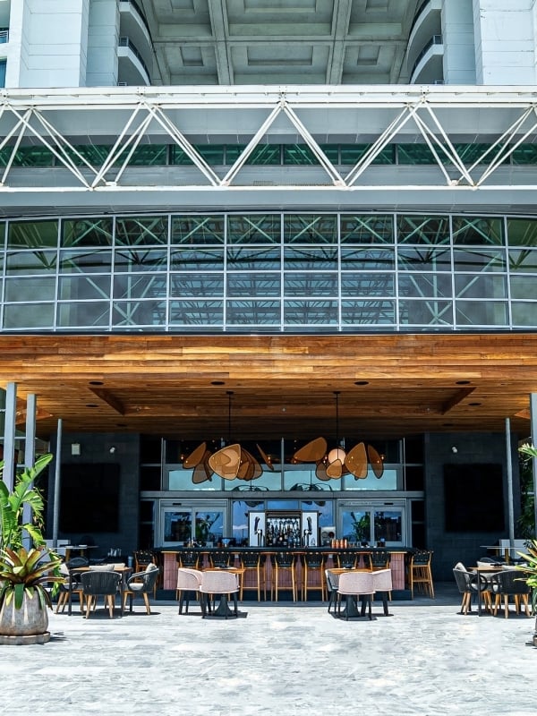 A modern outdoor bar and lounge area is situated beneath a multi-story glass facade, with a high-rise building visible above, connected by a white truss bridge.