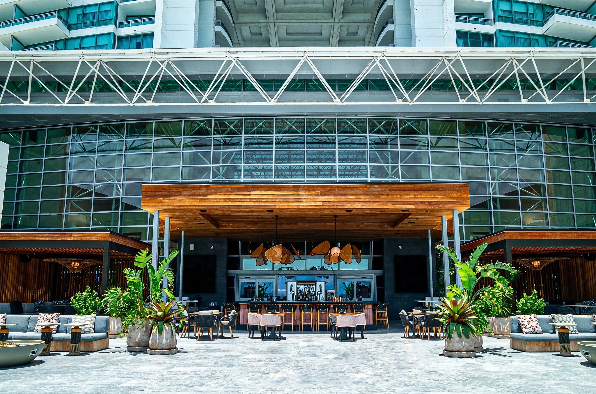 A modern outdoor bar and lounge area is situated beneath a multi-story glass facade, with a high-rise building visible above, connected by a white truss bridge.