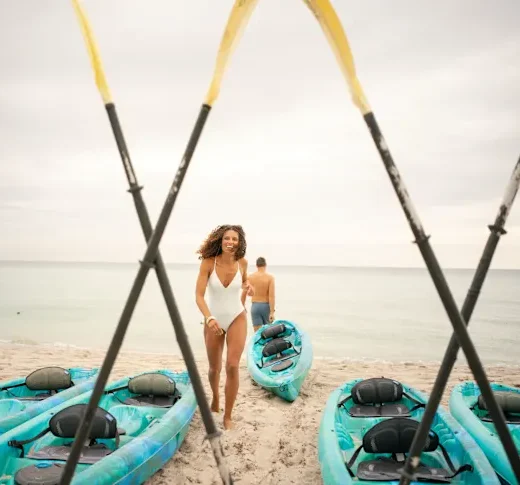 A couple preparing to kayak on the ocean. The man heads towards the ocean, kayak in tow, while the woman heads back to grab her kayak.