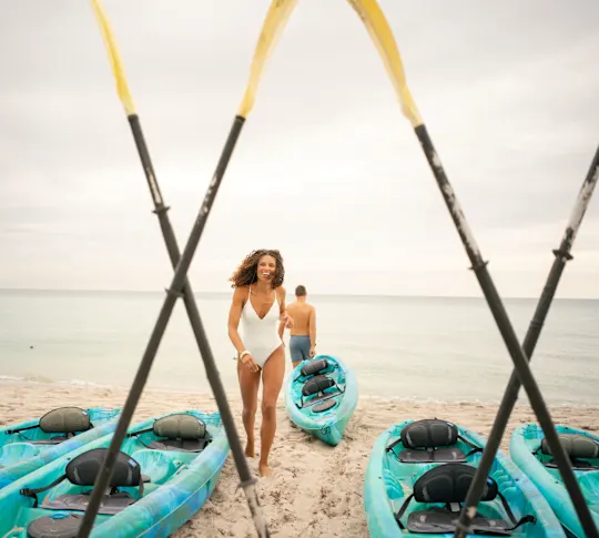 A couple preparing to kayak on the ocean. The man heads towards the ocean, kayak in tow, while the woman heads back to grab her kayak.
