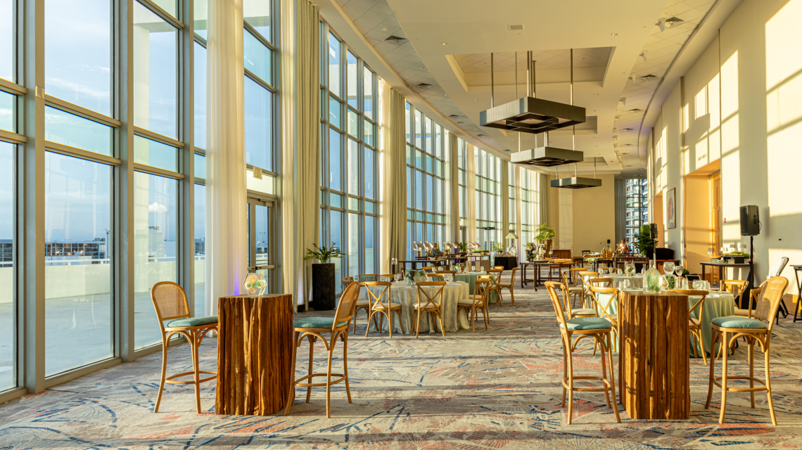 A bright, spacious event room is set up with dining and high-top tables. Sunlight streams through floor-to-ceiling windows overlooking an urban area. A sign reads "ROOM 2-3".