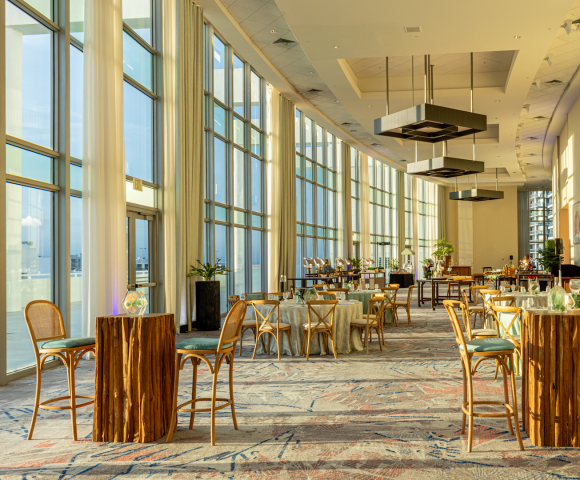 A bright, spacious event room is set up with dining and high-top tables. Sunlight streams through floor-to-ceiling windows overlooking an urban area. A sign reads "ROOM 2-3".