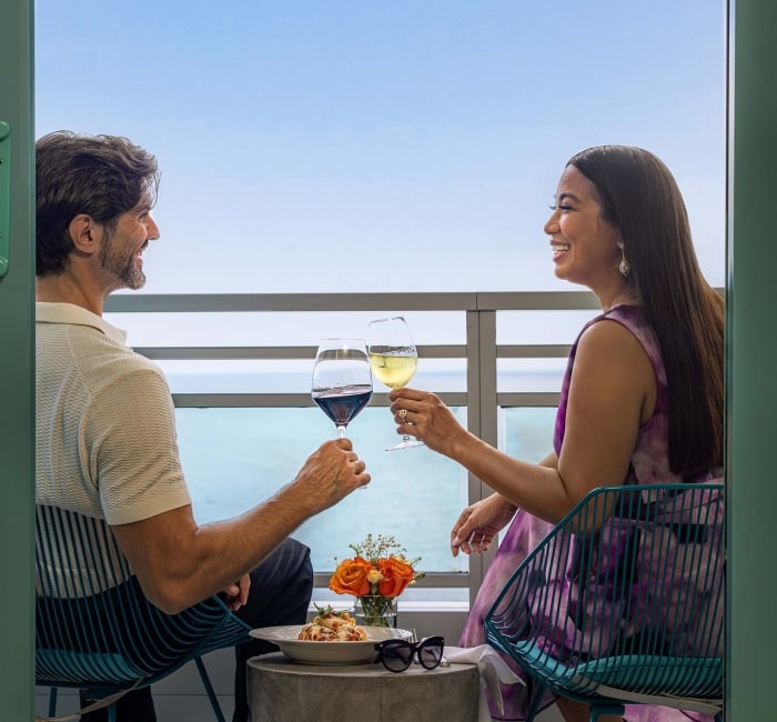 A smiling man and woman toast wine glasses across a small table with food and flowers, seated on a balcony overlooking the ocean.