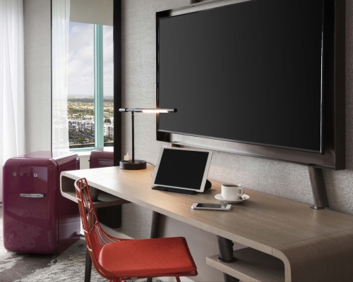 A contemporary desk setup includes a wall-mounted monitor, tablet, phone, and coffee cup. An orange chair and a red "SMEG" mini-fridge sit nearby, overlooking a city skyline.