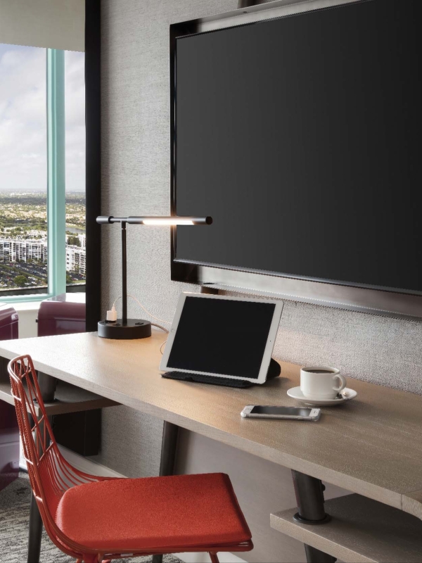 A contemporary desk setup includes a wall-mounted monitor, tablet, phone, and coffee cup. An orange chair and a red "SMEG" mini-fridge sit nearby, overlooking a city skyline.