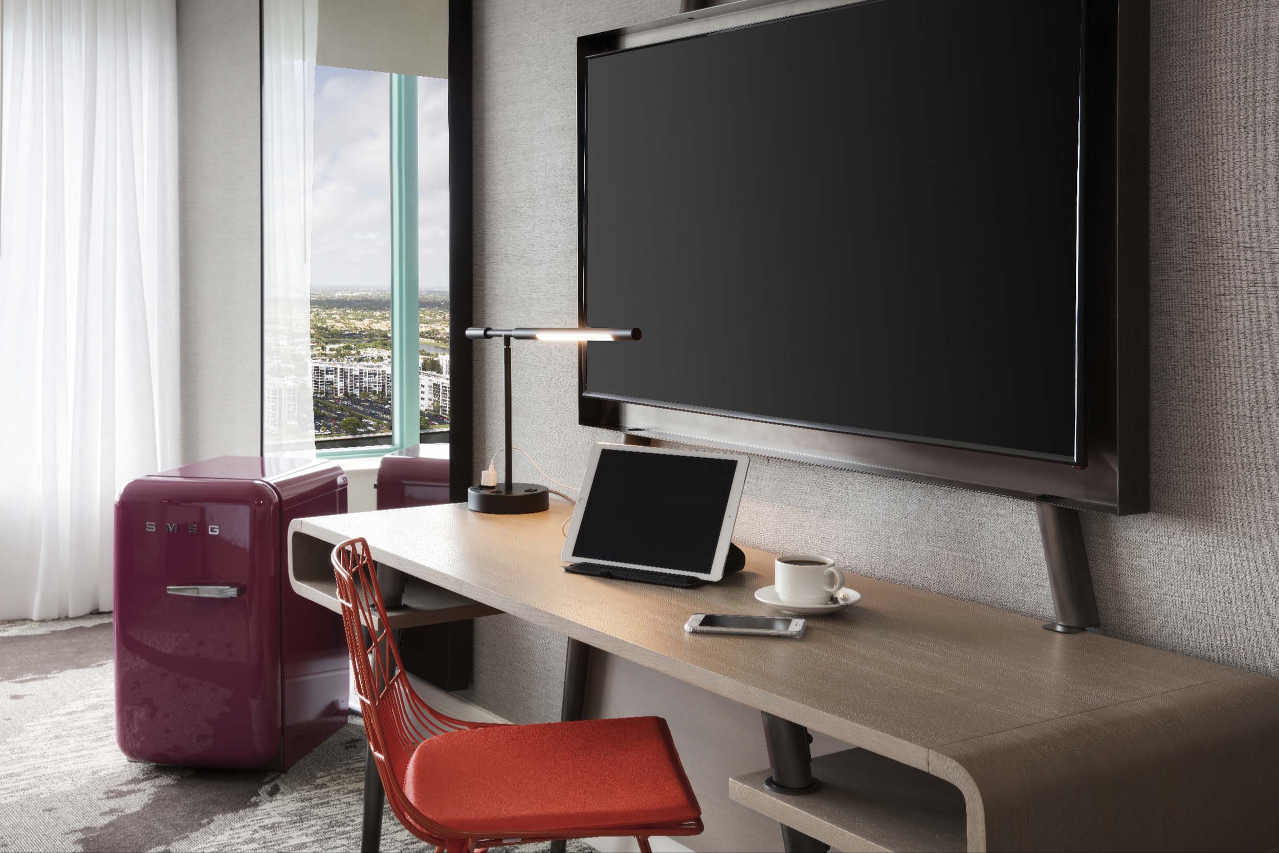 A contemporary desk setup includes a wall-mounted monitor, tablet, phone, and coffee cup. An orange chair and a red "SMEG" mini-fridge sit nearby, overlooking a city skyline.