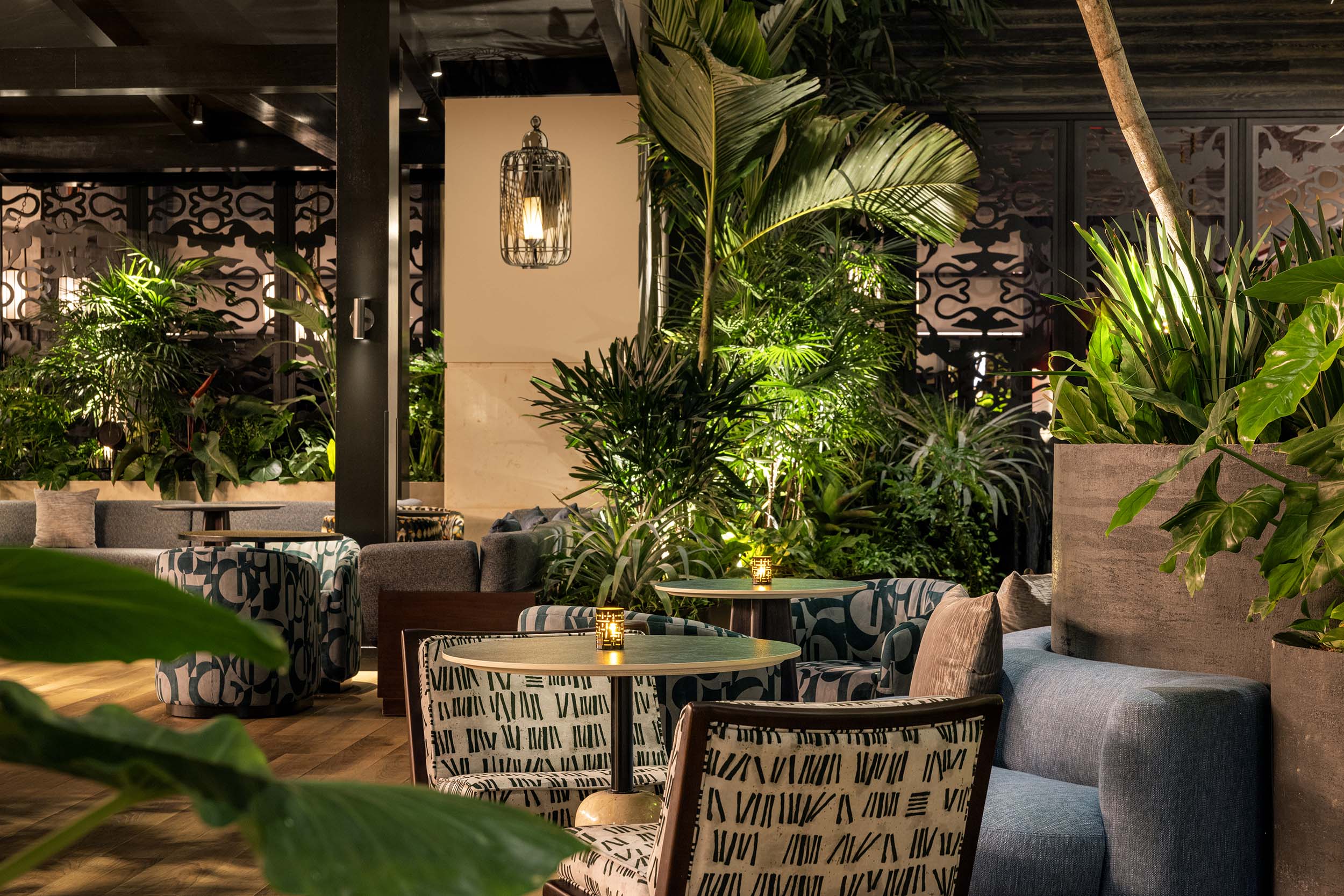 Restaurant interior featuring round tables with small lamps, varied seating, and lush green plants. Warm lighting illuminates the space, which includes dark ornate screens and wooden floors.