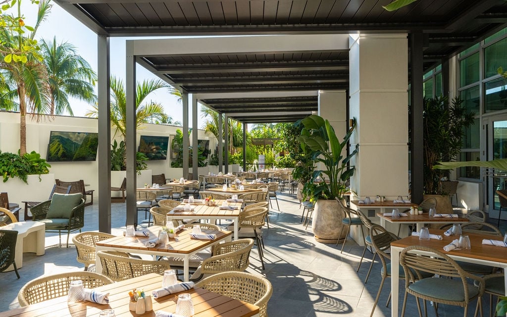 An outdoor restaurant patio features numerous rattan tables and chairs set for dining under a modern pergola. Lush palm trees and tropical plants adorn the bright, open-air space with wall-mounted TVs.
