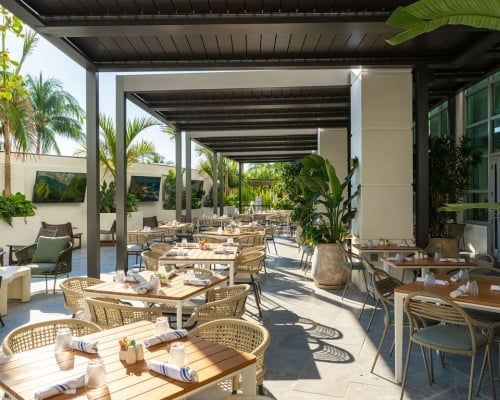 An outdoor restaurant patio features numerous rattan tables and chairs set for dining under a modern pergola. Lush palm trees and tropical plants adorn the bright, open-air space with wall-mounted TVs.