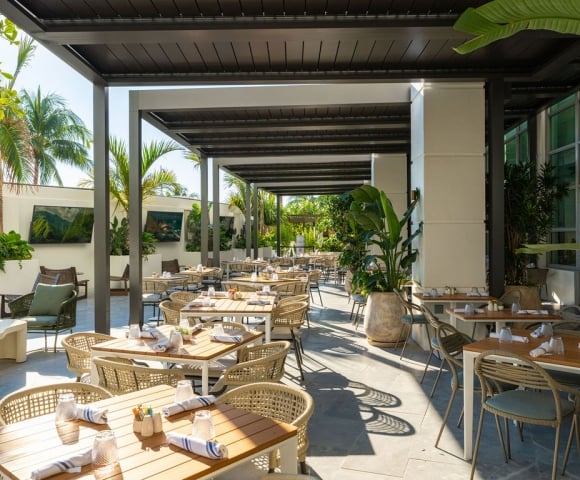 An outdoor restaurant patio features numerous rattan tables and chairs set for dining under a modern pergola. Lush palm trees and tropical plants adorn the bright, open-air space with wall-mounted TVs.