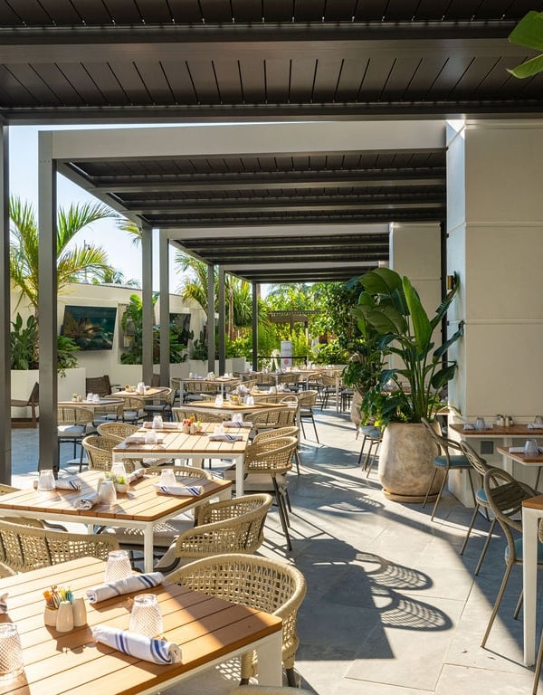 An outdoor restaurant patio features numerous rattan tables and chairs set for dining under a modern pergola. Lush palm trees and tropical plants adorn the bright, open-air space with wall-mounted TVs.