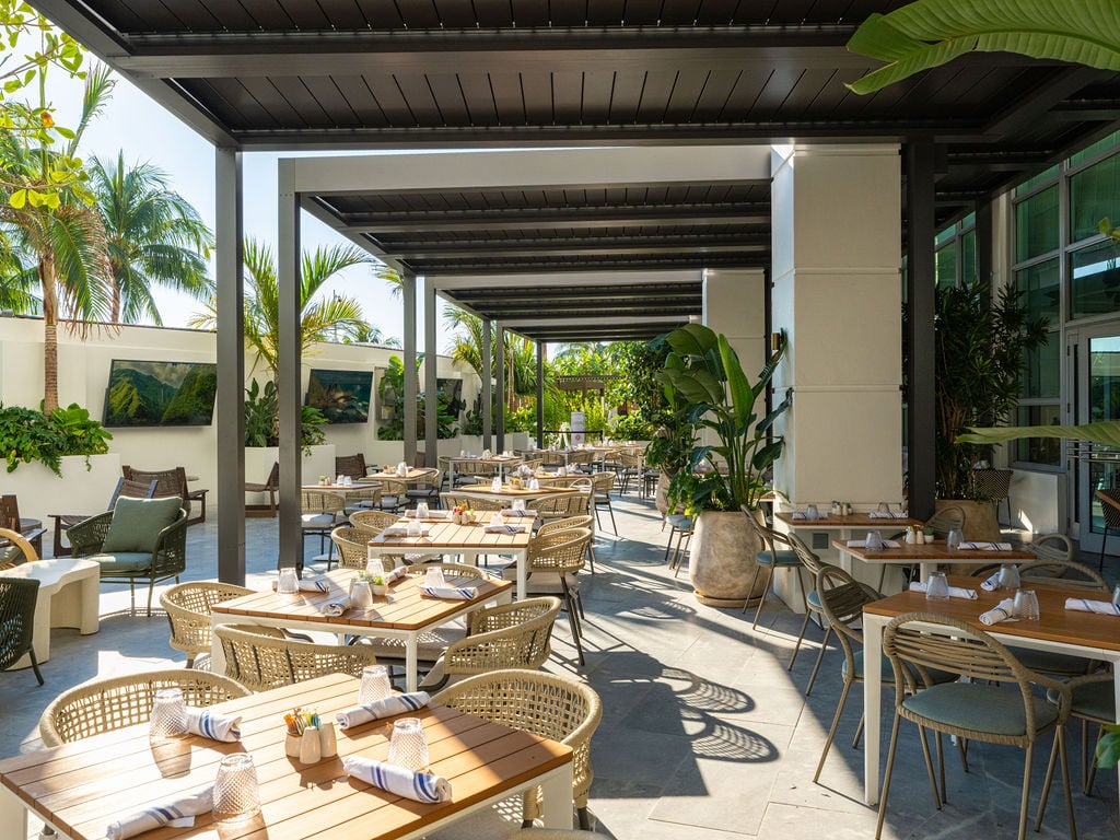 An outdoor restaurant patio features numerous rattan tables and chairs set for dining under a modern pergola. Lush palm trees and tropical plants adorn the bright, open-air space with wall-mounted TVs.