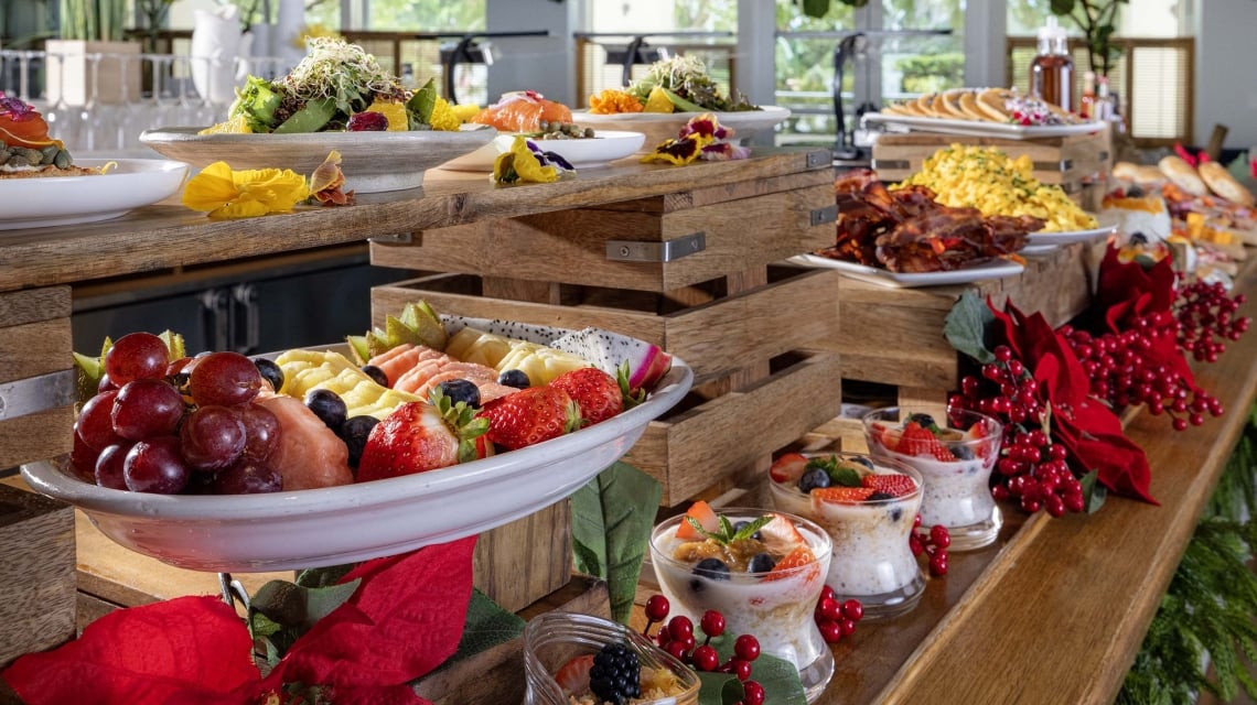 A lavish breakfast buffet displays various foods—fruit, pancakes, bacon, eggs, salmon, parfaits—on tiered wooden stands with red festive décor in a bright room.