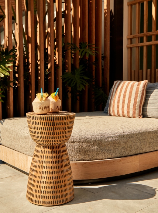 A luxury poolside daybed with pillows next to a woven side table holding two tropical coconut drinks.