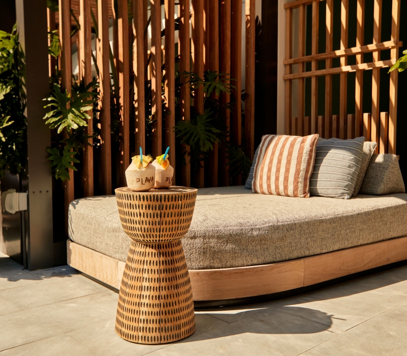 A luxury poolside daybed with pillows next to a woven side table holding two tropical coconut drinks.