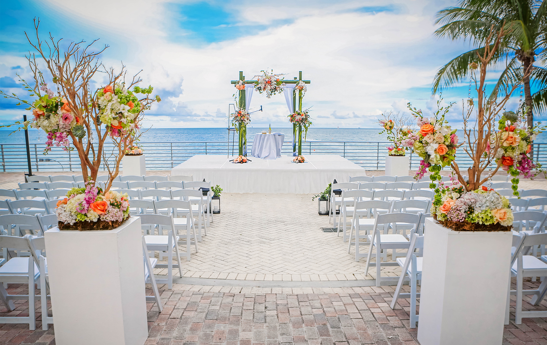 A wedding ceremony setup features a floral arch on a stage with white chairs on a brick patio. Floral pedestals line the aisle, overlooking the ocean under a cloudy sky.