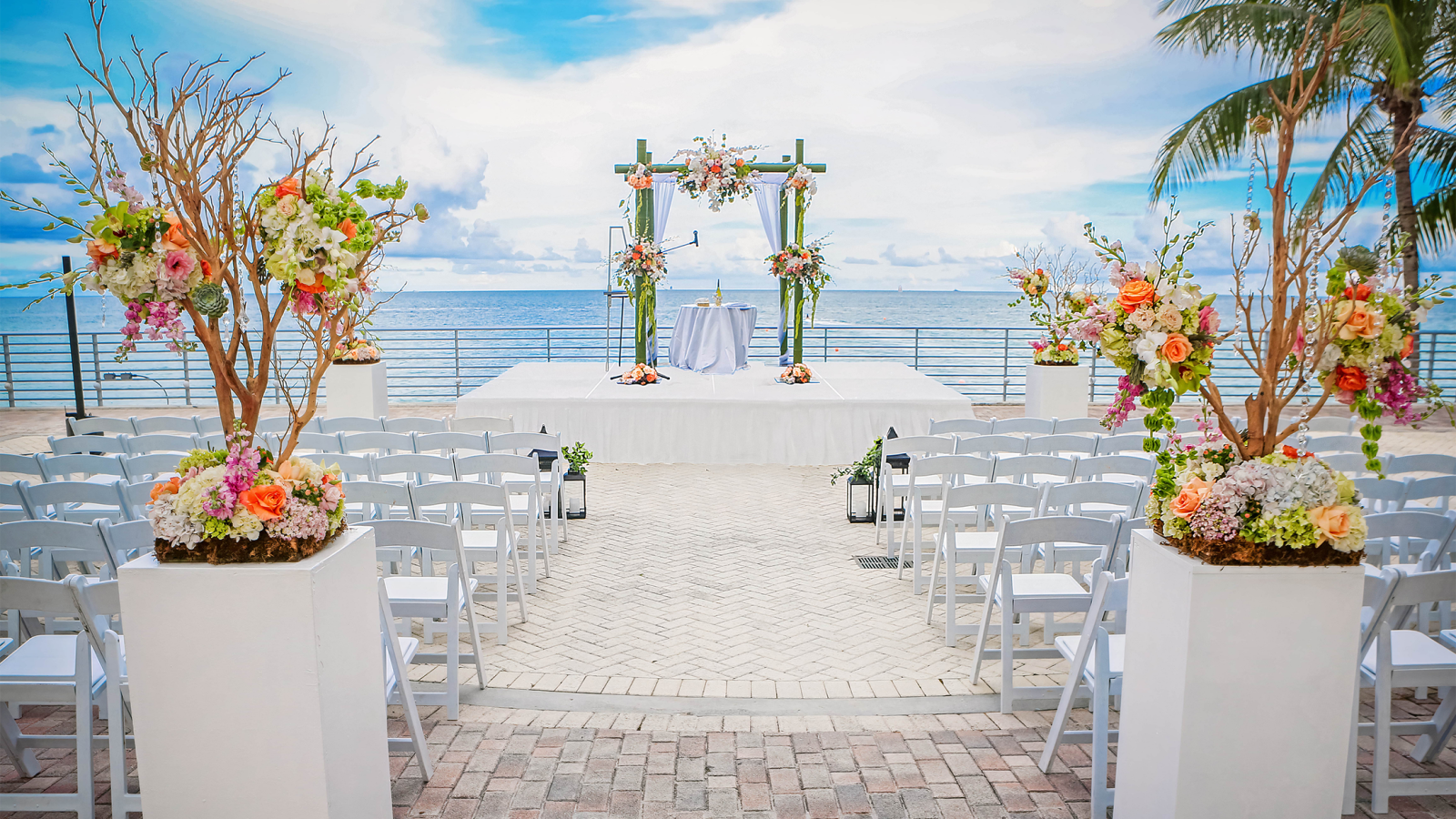 A wedding ceremony setup features a floral arch on a stage with white chairs on a brick patio. Floral pedestals line the aisle, overlooking the ocean under a cloudy sky.
