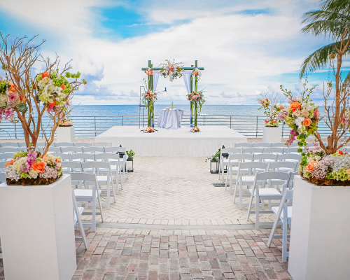 A wedding ceremony setup features a floral arch on a stage with white chairs on a brick patio. Floral pedestals line the aisle, overlooking the ocean under a cloudy sky.