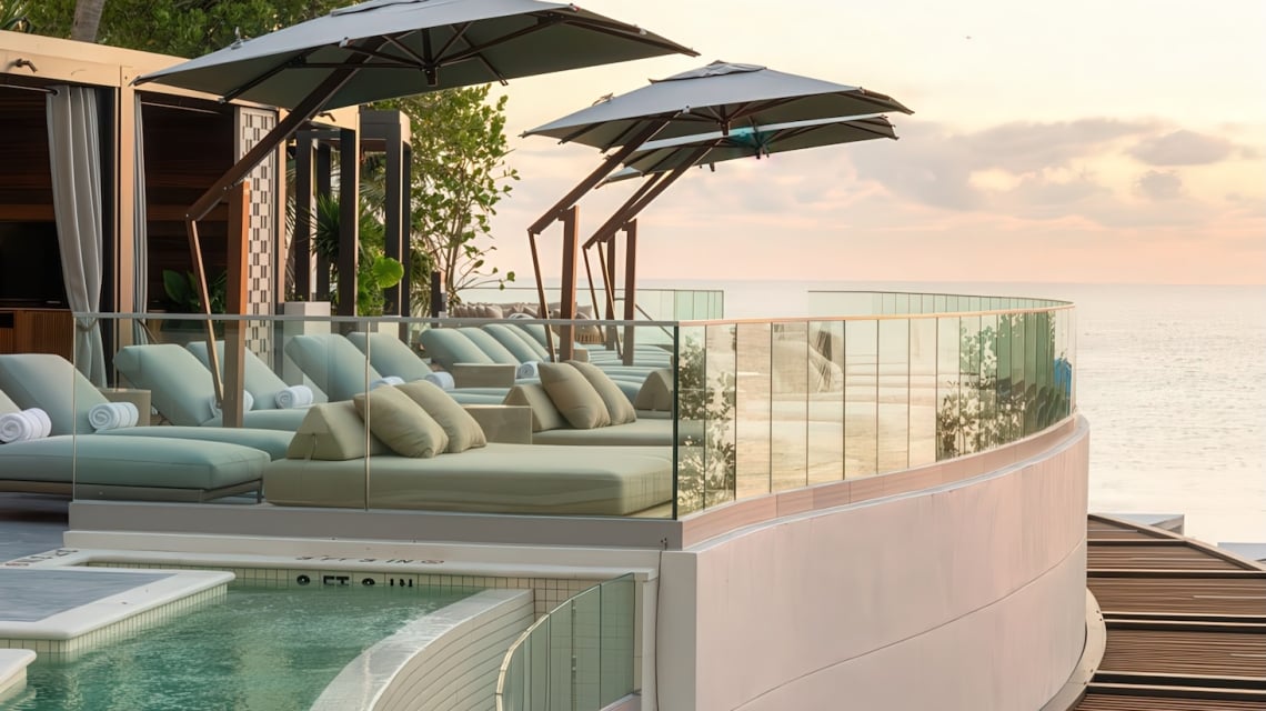 Resort sun lounge and pool deck features multiple daybeds under umbrellas arranged along a curved pool (marked 2 FT, 3 FT, 4 FT) and glass balcony, overlooking the calm ocean at sunset, with palm trees visible.