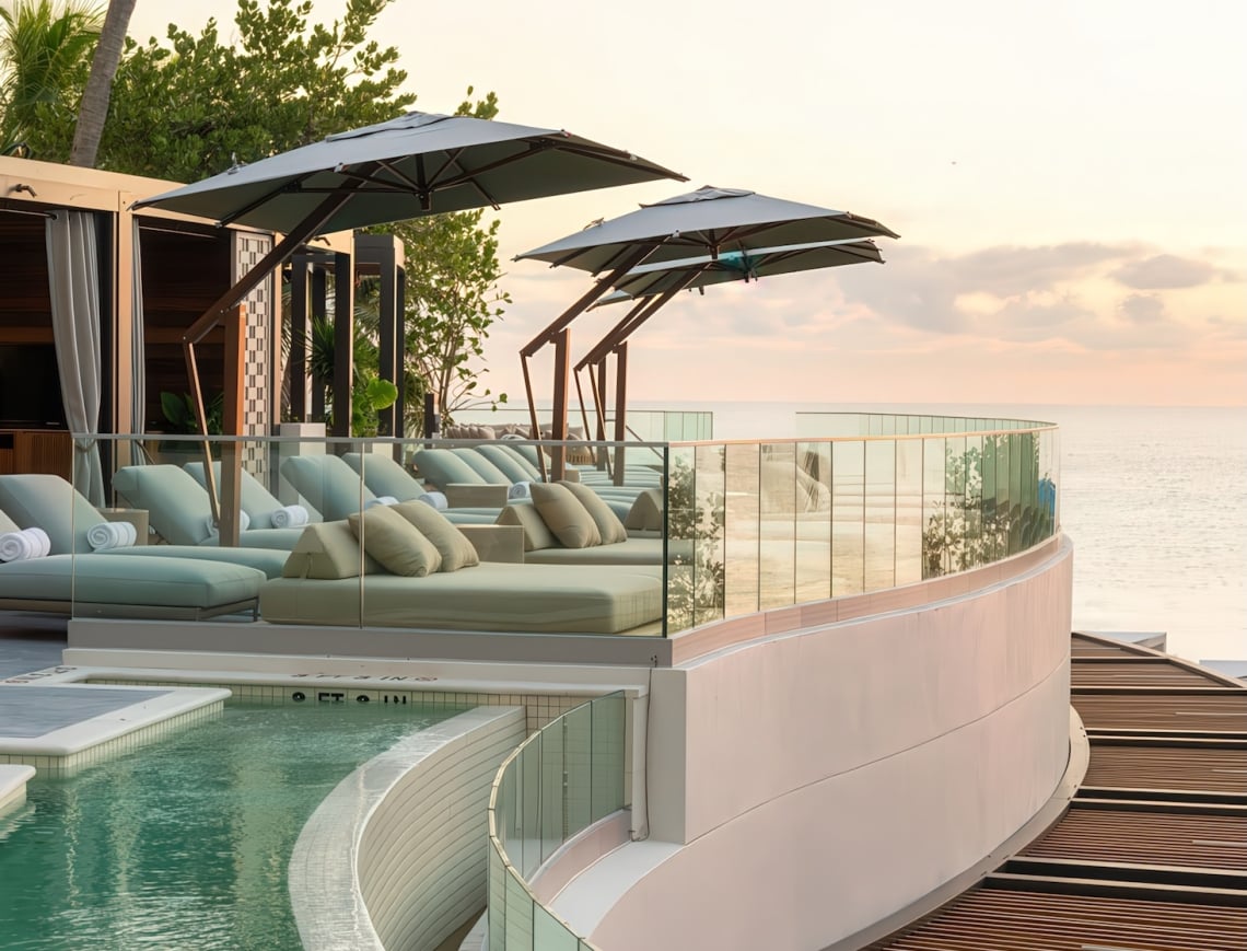 Resort sun lounge and pool deck features multiple daybeds under umbrellas arranged along a curved pool (marked 2 FT, 3 FT, 4 FT) and glass balcony, overlooking the calm ocean at sunset, with palm trees visible.