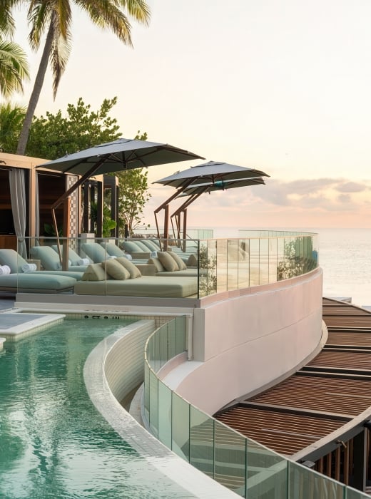 Resort sun lounge and pool deck features multiple daybeds under umbrellas arranged along a curved pool (marked 2 FT, 3 FT, 4 FT) and glass balcony, overlooking the calm ocean at sunset, with palm trees visible.