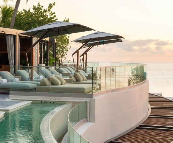 Resort sun lounge and pool deck features multiple daybeds under umbrellas arranged along a curved pool (marked 2 FT, 3 FT, 4 FT) and glass balcony, overlooking the calm ocean at sunset, with palm trees visible.