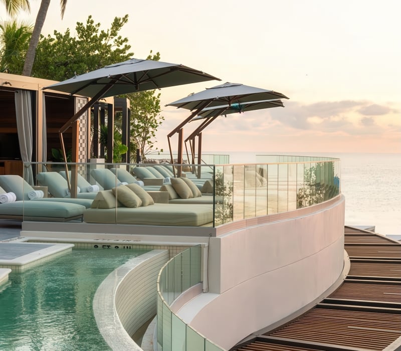 Resort sun lounge and pool deck features multiple daybeds under umbrellas arranged along a curved pool (marked 2 FT, 3 FT, 4 FT) and glass balcony, overlooking the calm ocean at sunset, with palm trees visible.