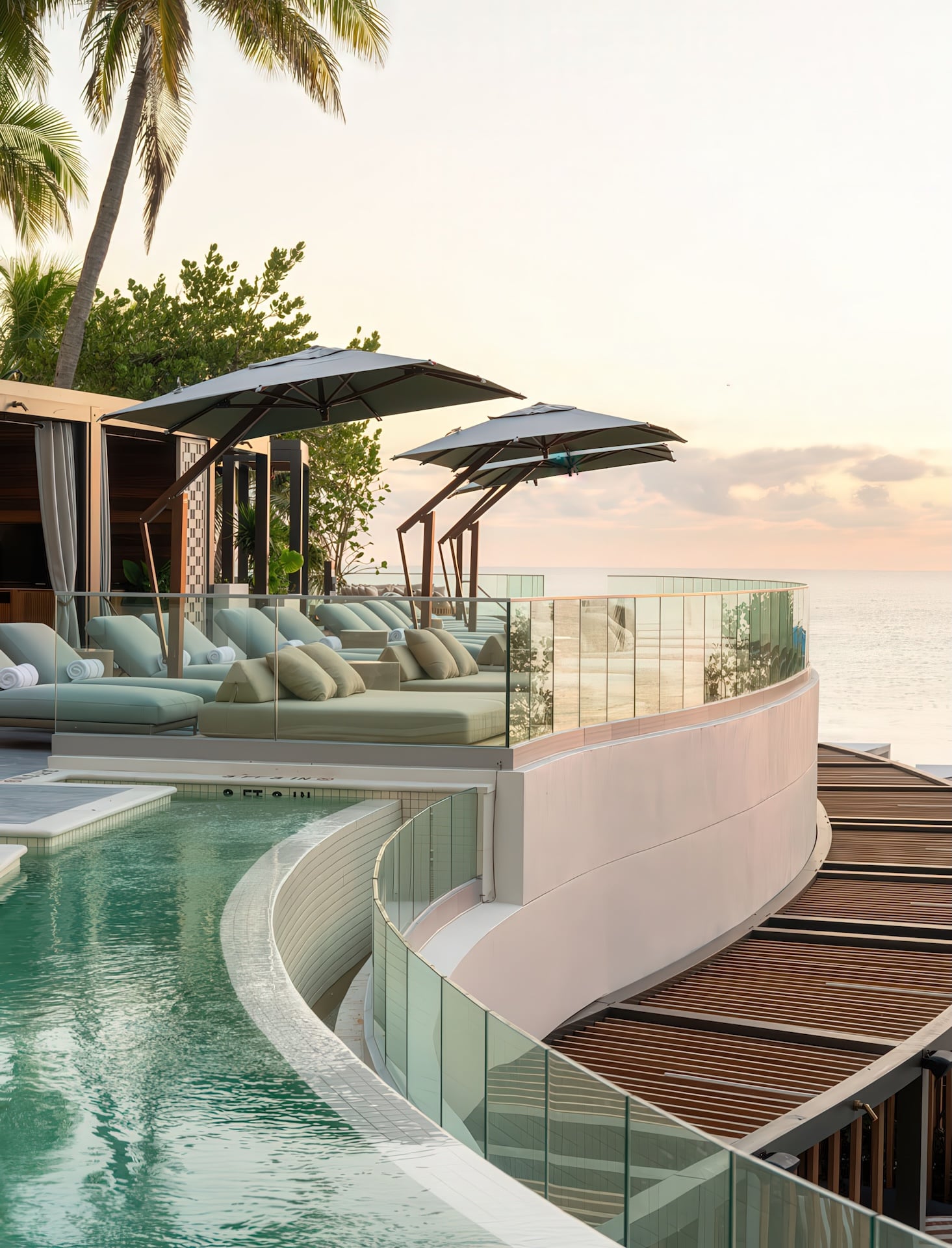 Resort sun lounge and pool deck features multiple daybeds under umbrellas arranged along a curved pool (marked 2 FT, 3 FT, 4 FT) and glass balcony, overlooking the calm ocean at sunset, with palm trees visible.