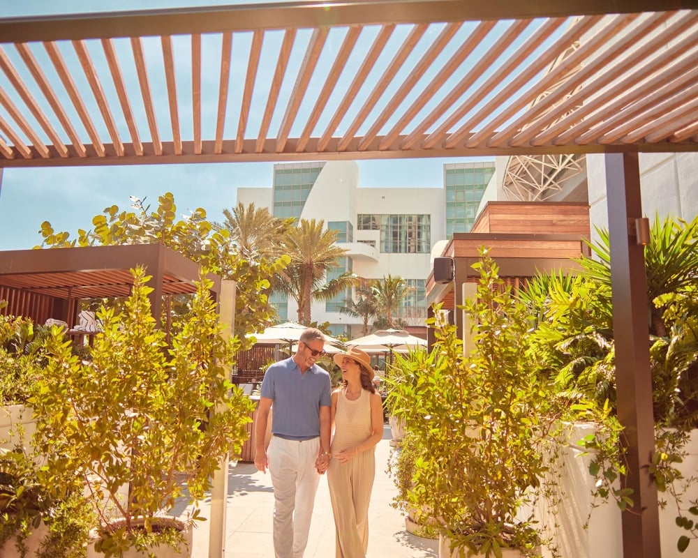 A couple holding hands walk under a wooden pergola surrounded by green plants