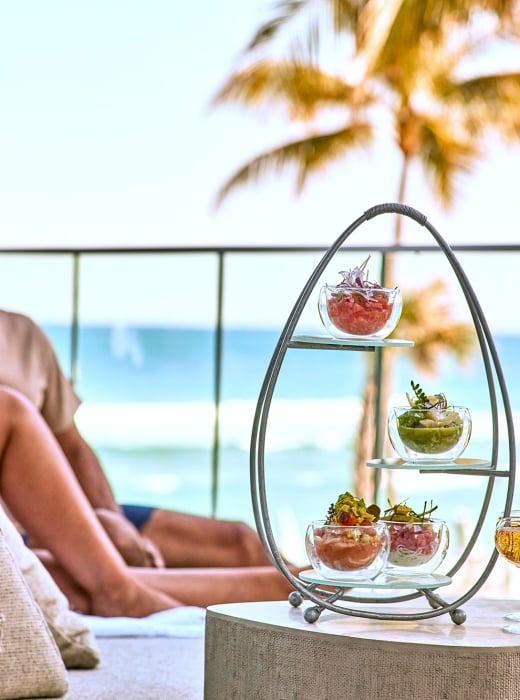 A tiered oval serving stand with small dishes and a wine glass on an oceanfront terrace, with a relaxed couple and palm trees behind.
