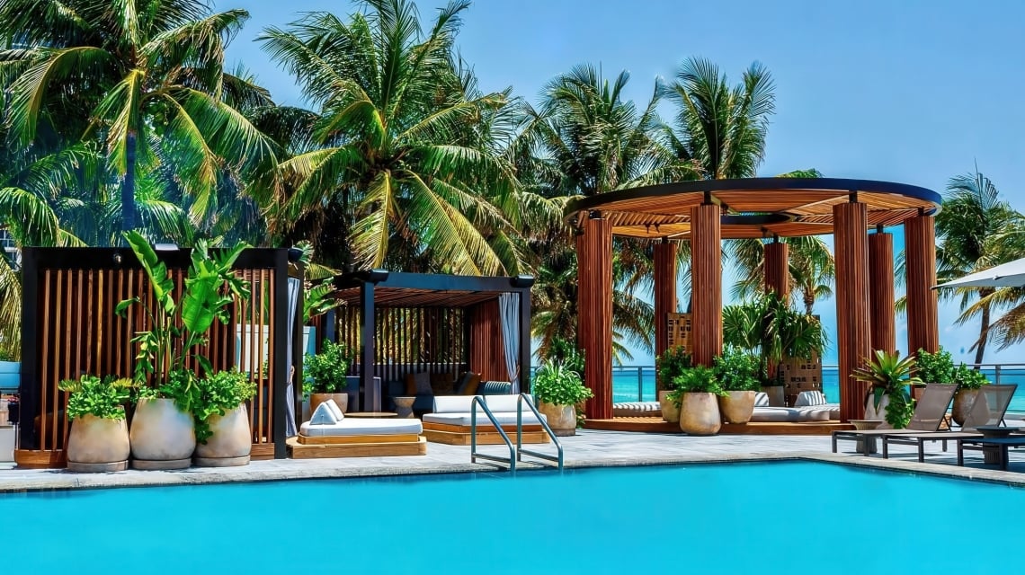A bright blue swimming pool is surrounded by wooden pergolas with lounge beds and potted plants, under tall palm trees and a clear sky, with the ocean visible in the background.