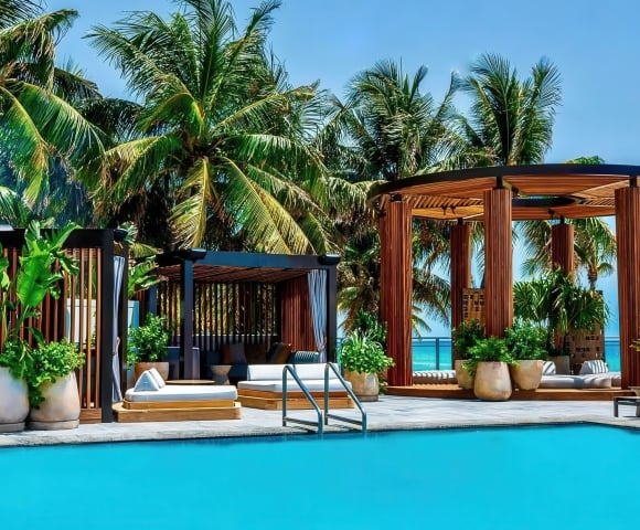 A bright blue swimming pool is surrounded by wooden pergolas with lounge beds and potted plants, under tall palm trees and a clear sky, with the ocean visible in the background.