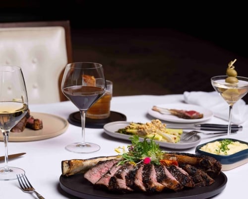 Sliced tomahawk steak, garnished, rests on a dark plate, flanked by white wine, red wine, and a martini, plus side dishes on a white-clothed table.