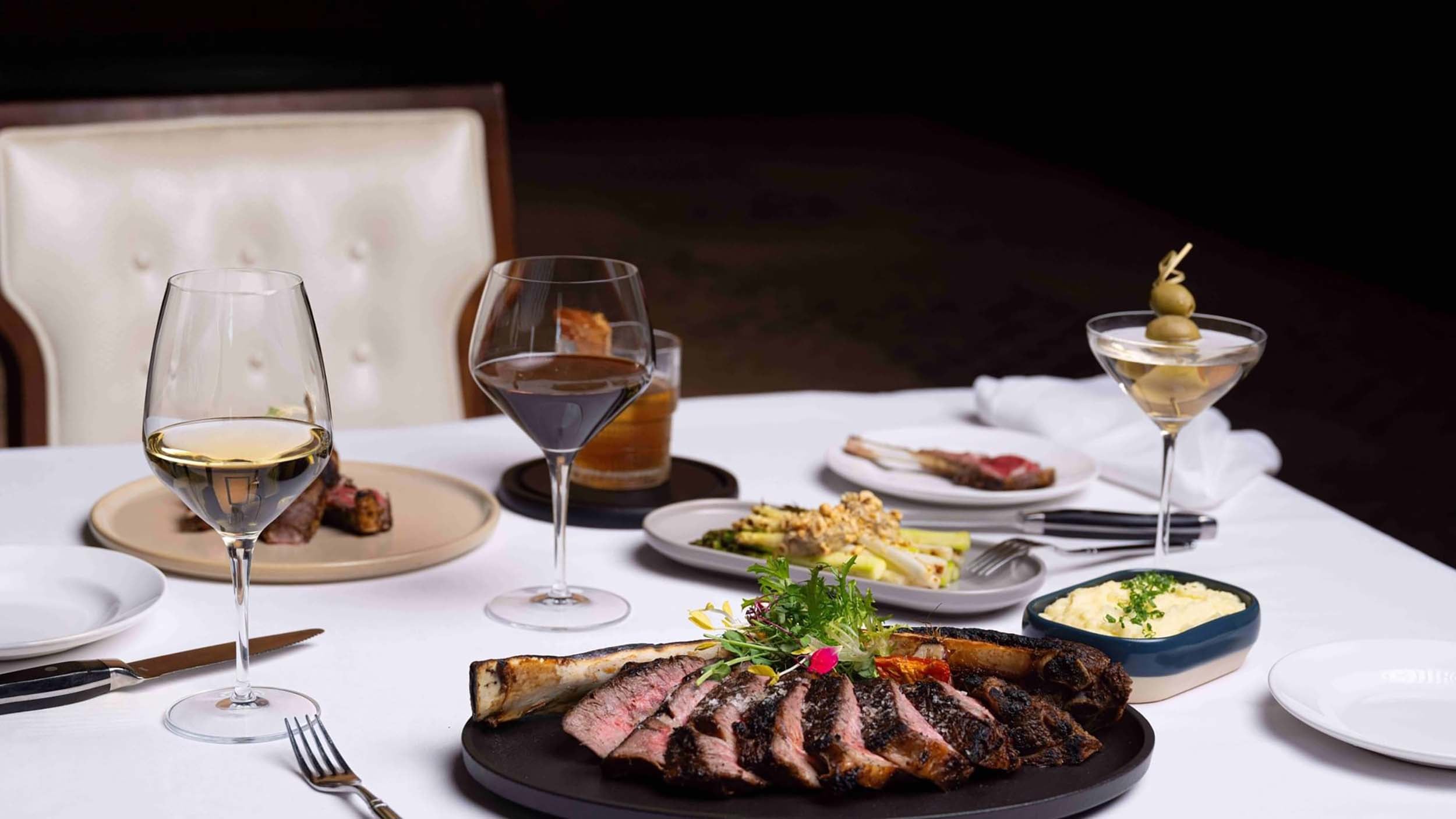 Sliced tomahawk steak, garnished, rests on a dark plate, flanked by white wine, red wine, and a martini, plus side dishes on a white-clothed table.