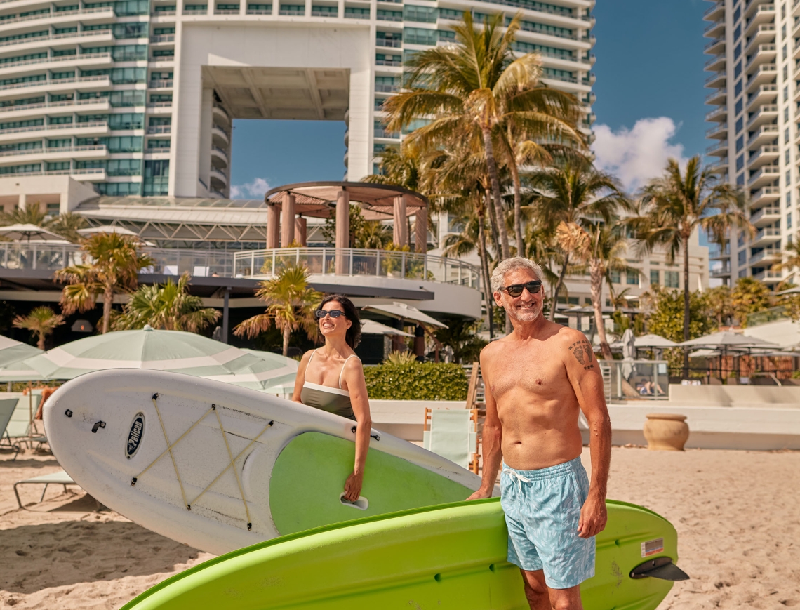 A couple on the beach in swim wear with paddleboards underarm