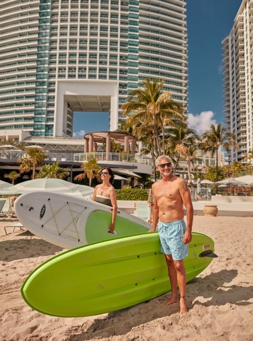 A couple on the beach in swim wear with paddleboards underarm