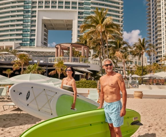A couple on the beach in swim wear with paddleboards underarm