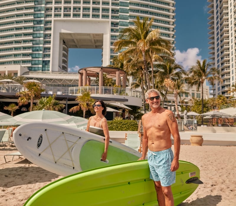 A couple on the beach in swim wear with paddleboards underarm