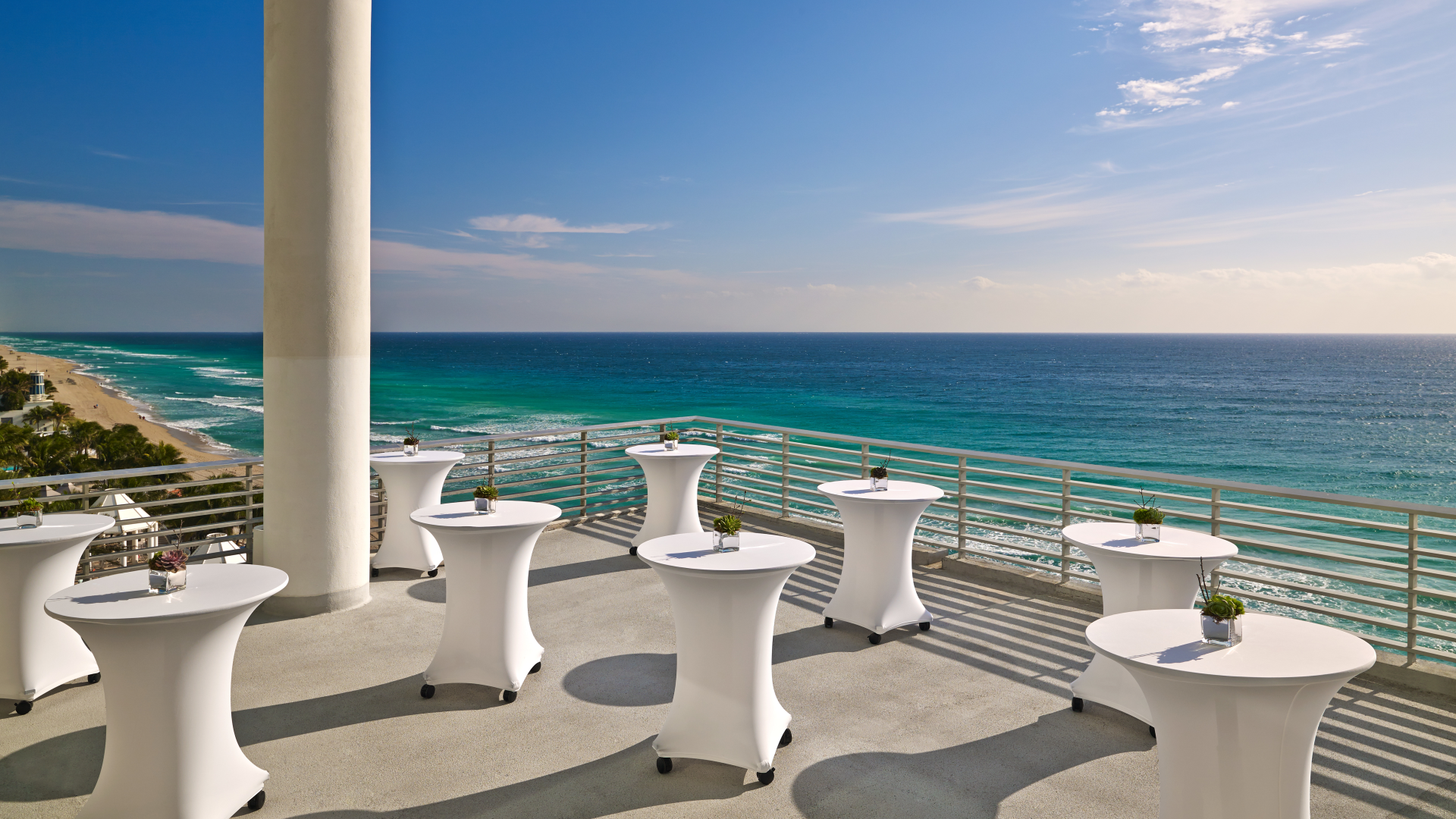 Several white high-top tables feature small succulent centerpieces on an outdoor terrace, overlooking the ocean and a distant sandy beach.
