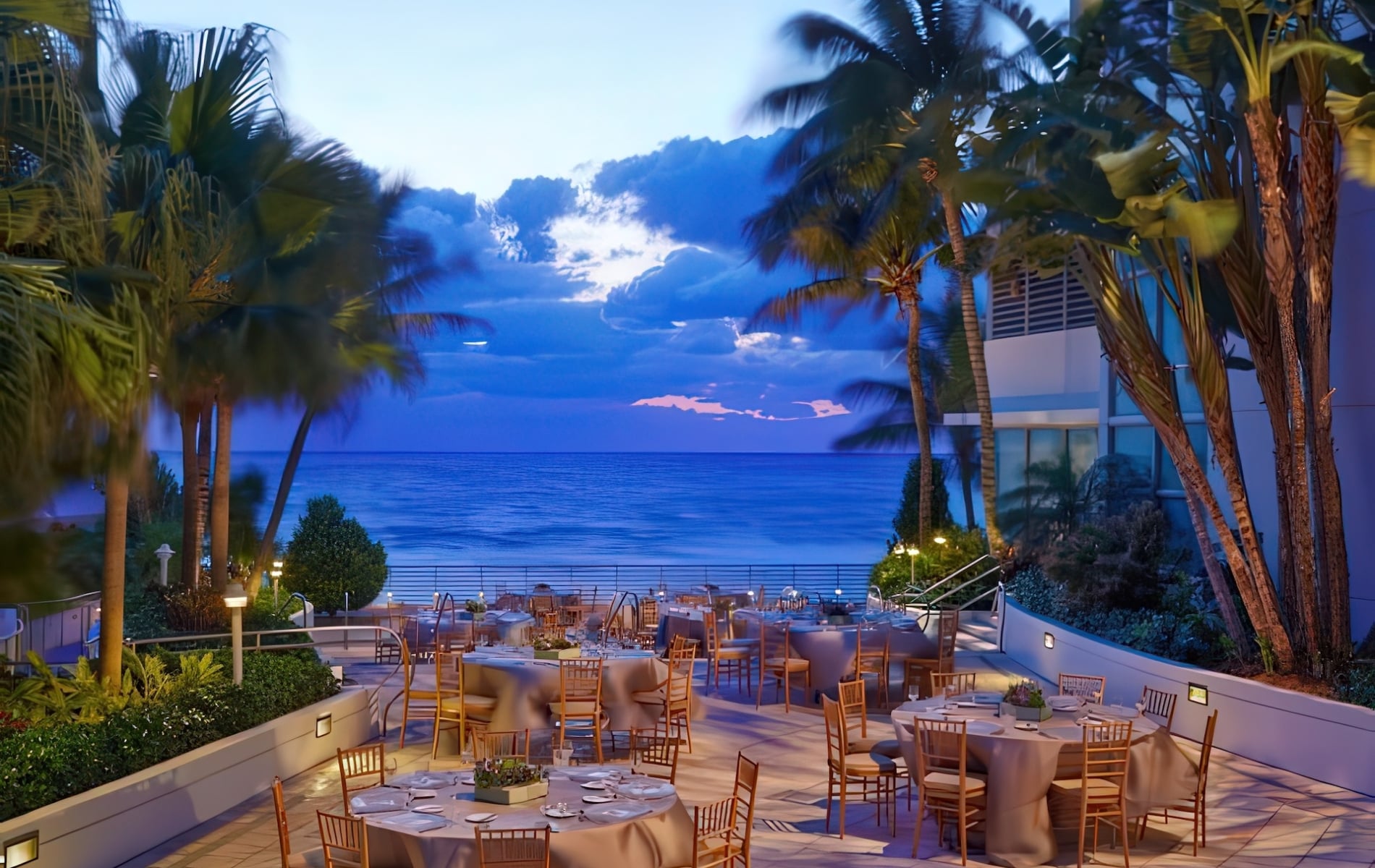 Set dining tables with chairs occupy an outdoor patio overlooking a tranquil blue ocean and twilight sky, surrounded by palm trees.