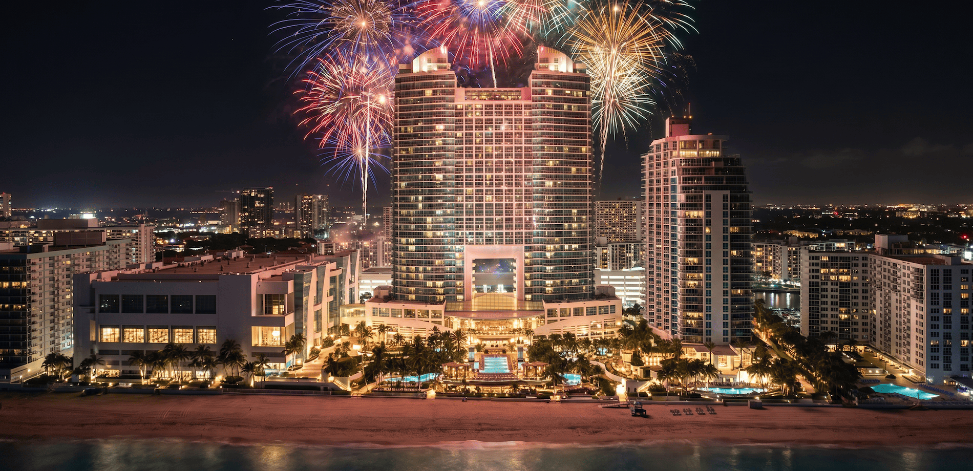 Colorful fireworks burst above a large, illuminated hotel complex on a beach, with city lights and the ocean at night.