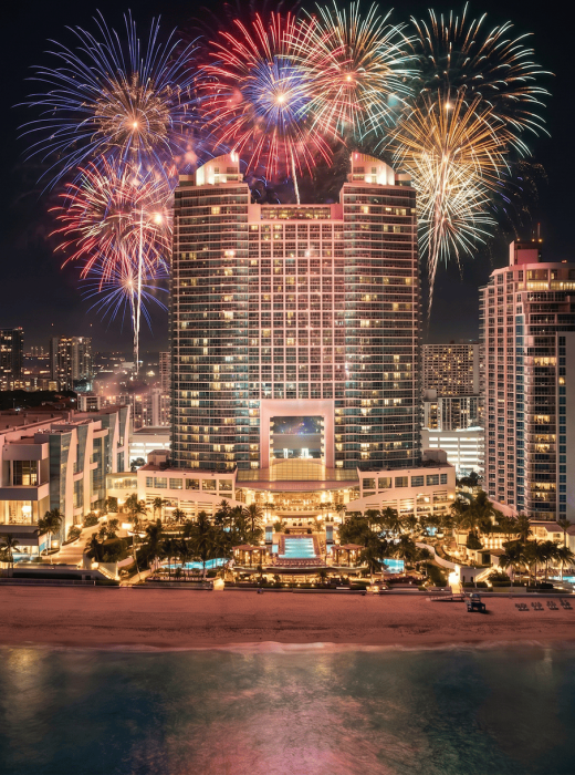 Colorful fireworks burst above a large, illuminated hotel complex on a beach, with city lights and the ocean at night.