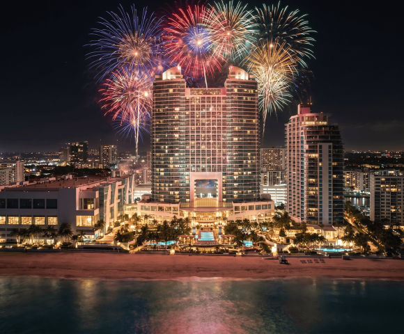 Colorful fireworks burst above a large, illuminated hotel complex on a beach, with city lights and the ocean at night.