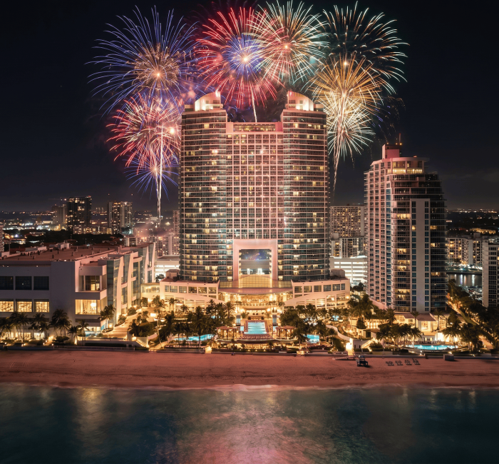 Colorful fireworks burst above a large, illuminated hotel complex on a beach, with city lights and the ocean at night.