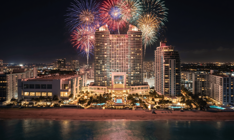 Colorful fireworks burst above a large, illuminated hotel complex on a beach, with city lights and the ocean at night.